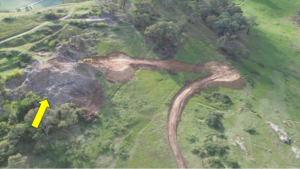 Aerial view of a hillside construction site with a curved dirt trench and a yellow excavator at work among trees and grass.