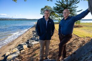 Two men stand on a rocky lakeshore under a clear blue sky, smiling at the camera.